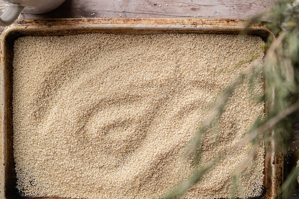 Overhead landscape view of sesame seeds spread in an even layer on a well-used baking sheet.