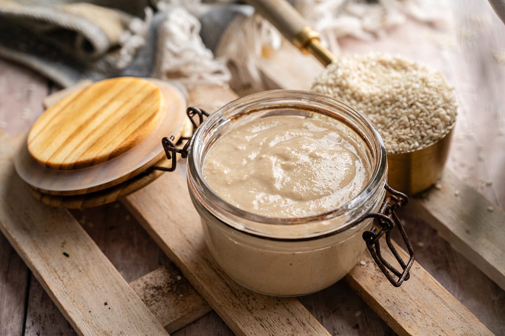 An open glass jar of homemade tahini beside its wooden lid and a measuring cup filled with sesame seeds.