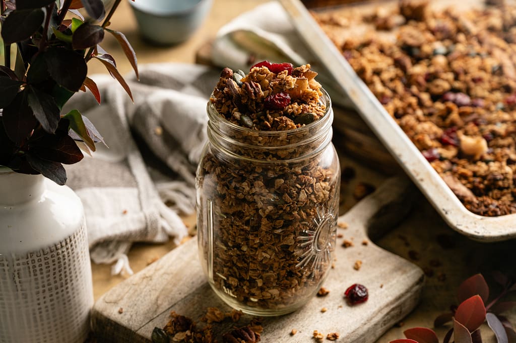 A glass jar of golden harvest granola beside a baking tray and brass measuring cup.