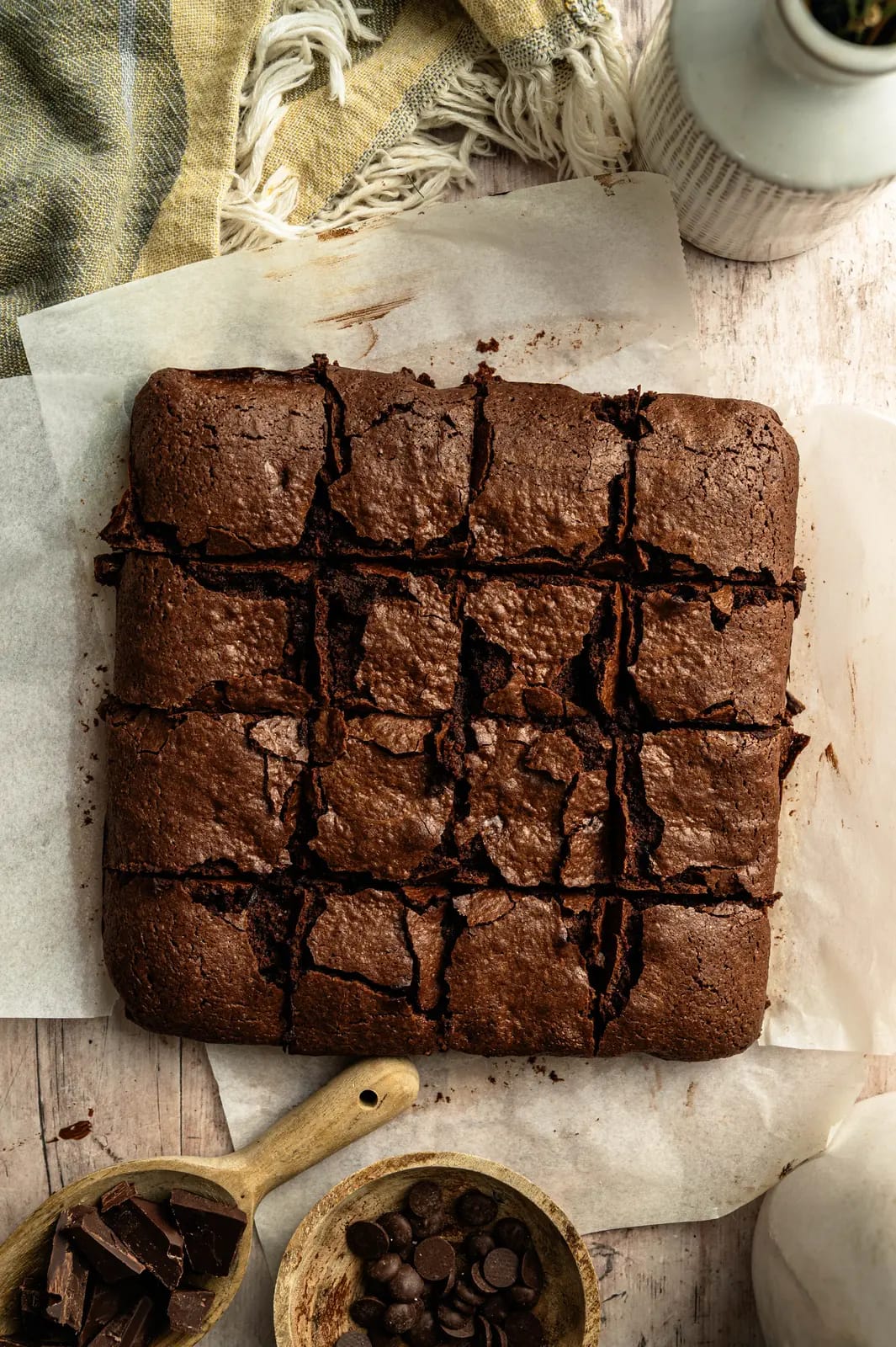 Overhead view of sixteen chocolate brownie squares on parchment beside chocolate chips.