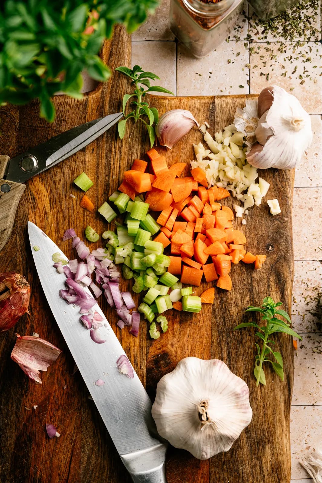 Chopped carrot, celery, onion, and garlic on a wooden board beside a knife.
