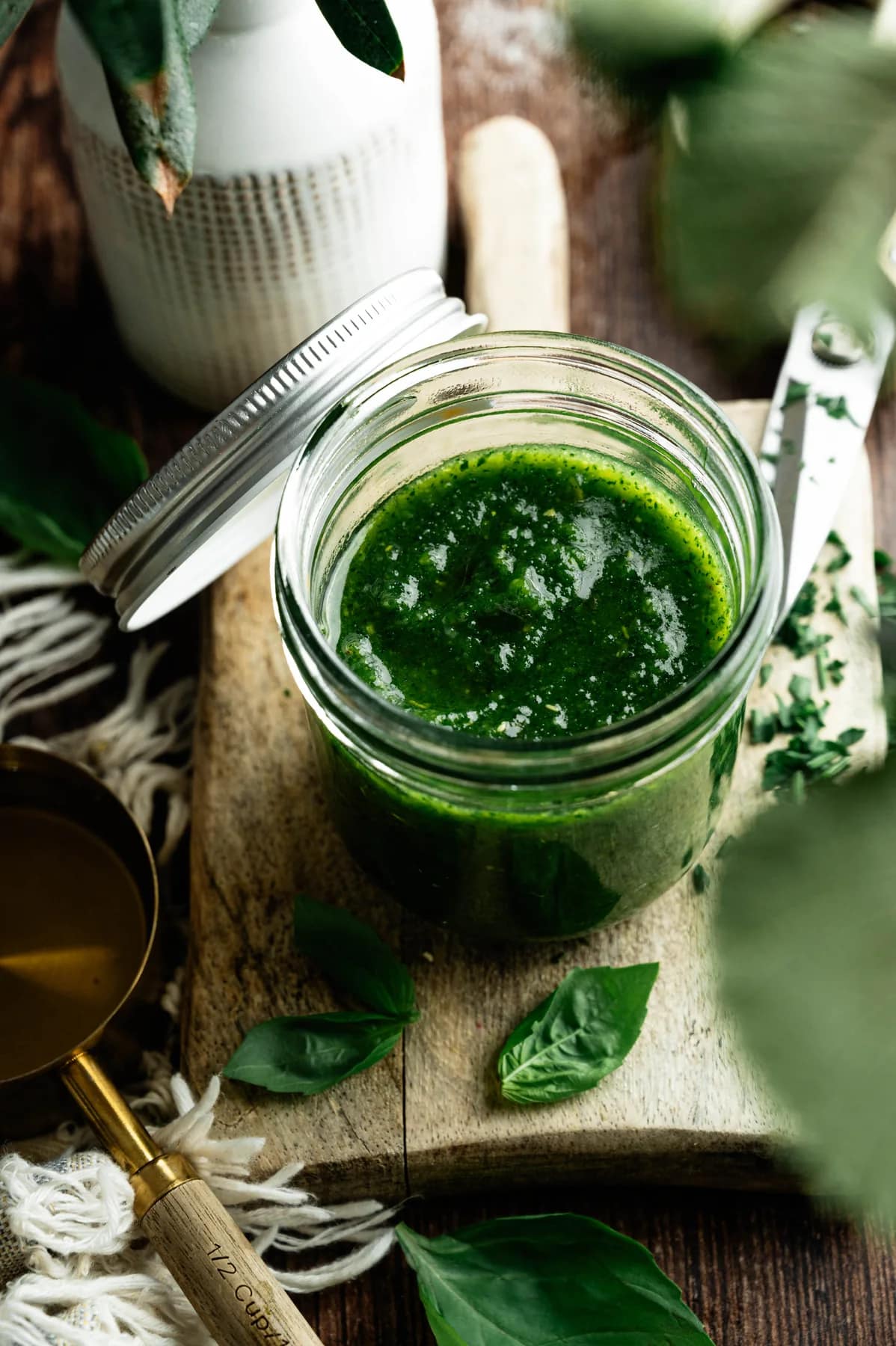 A jar of homemade basil pesto on a wooden board with fresh basil leaves.