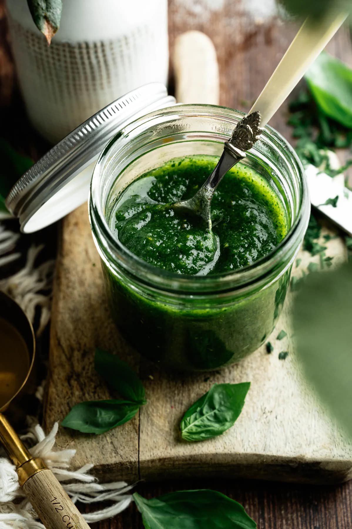 A spoon in a jar of basil pesto on a wooden board with fresh basil leaves.