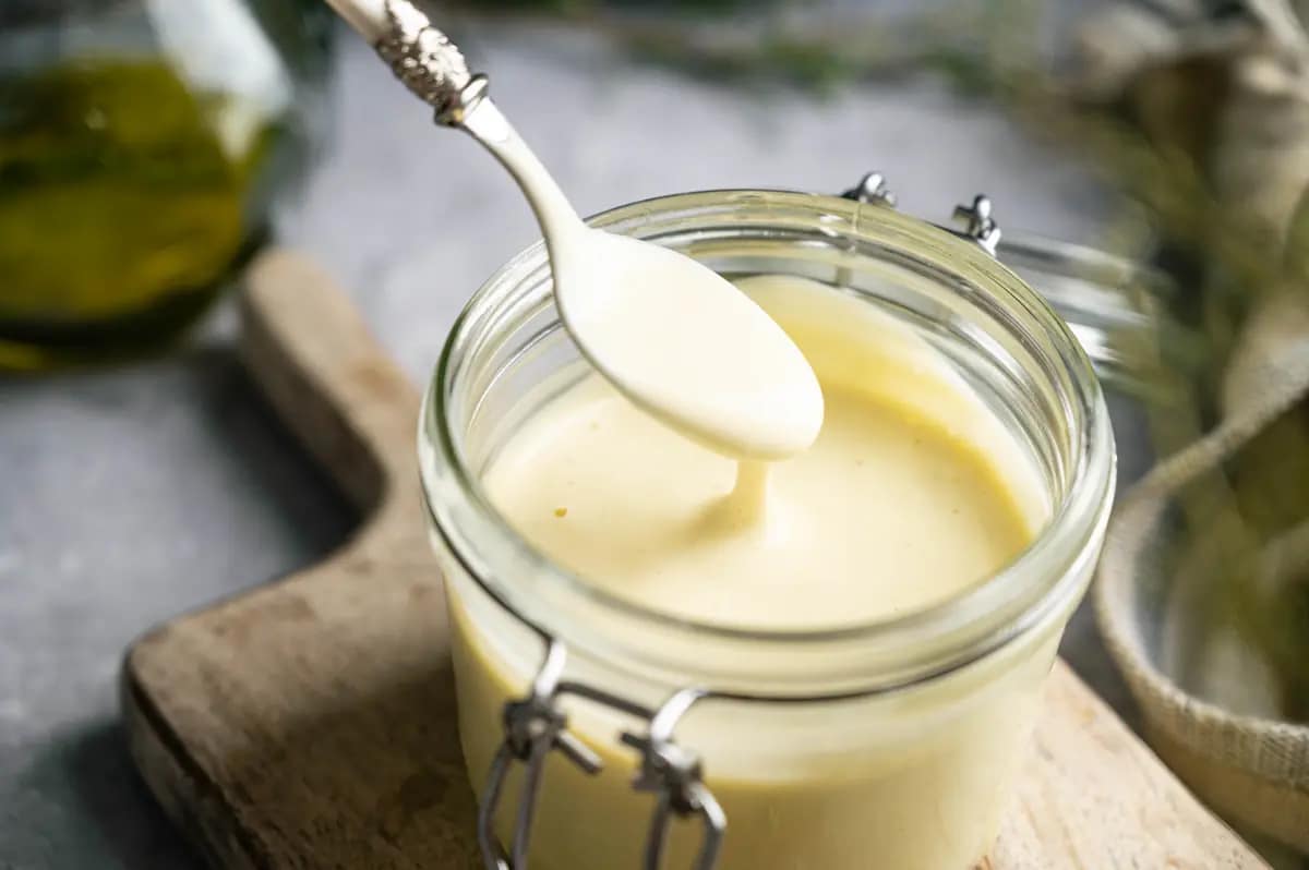 A spoon lifts creamy pale yellow mayonnaise from a glass clamp-top jar.
