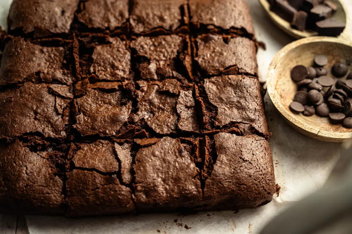 Chocolate brownie squares with crackled tops beside a bowl of chocolate chips.