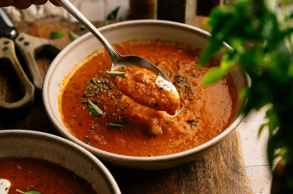 A spoon lifts thick tomato basil soup from a ceramic bowl.