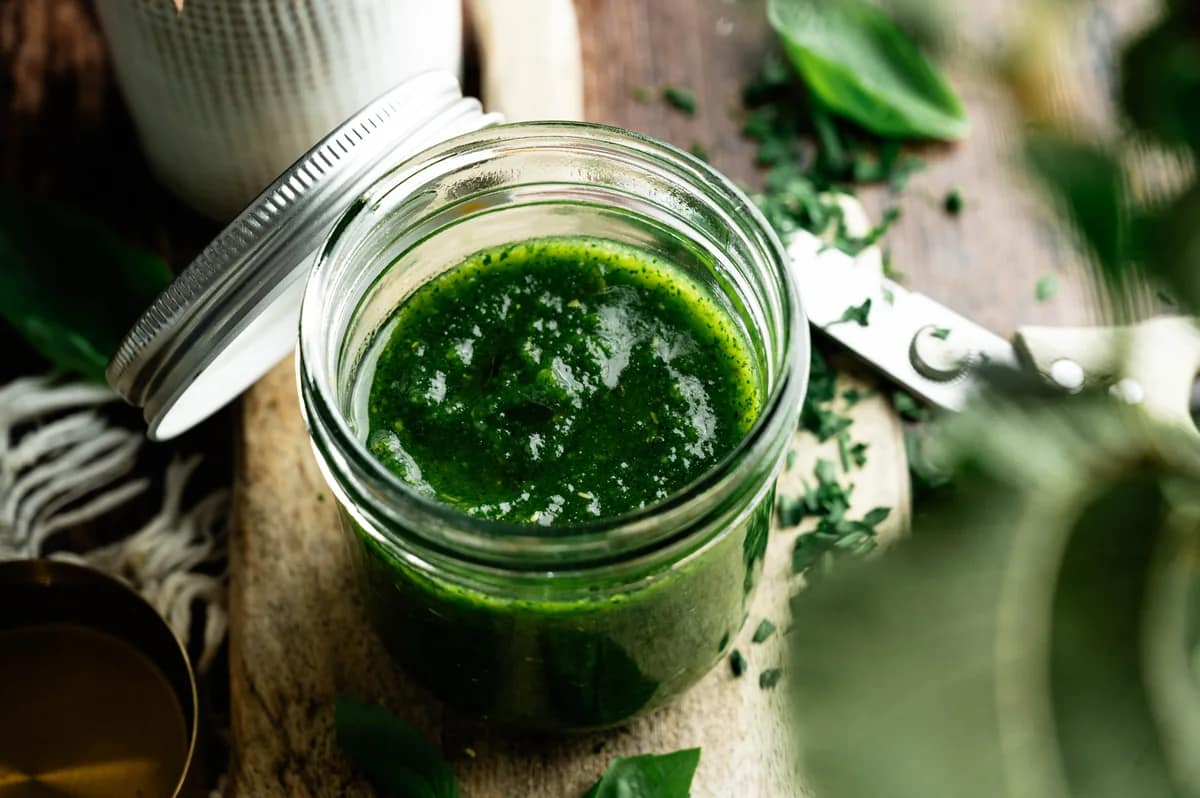 Horizontal view of a jar of basil pesto on a wooden board beside a white jug.