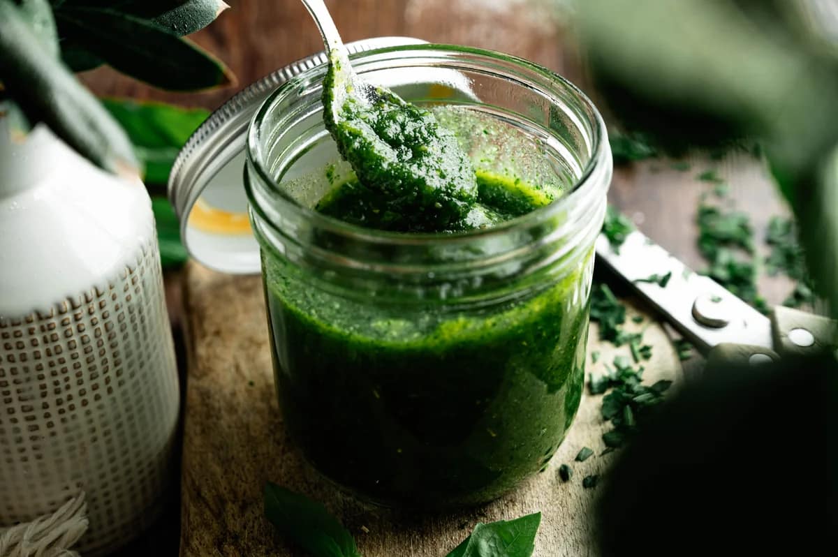 A spoonful of basil pesto held above an open jar on a rustic wooden table.