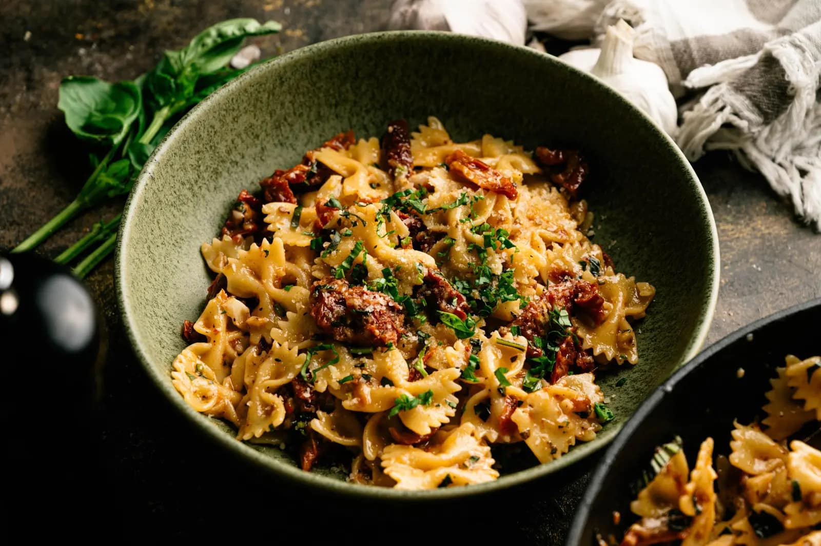 A green bowl of sun-dried tomato and basil farfalle with grated cheese, beside garlic and a skillet of pasta.