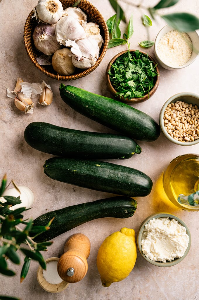 Ingredients for lemon ricotta zoodles arranged with zucchini, ricotta, lemon, herbs, garlic, Parmesan, pine nuts, and olive oil.