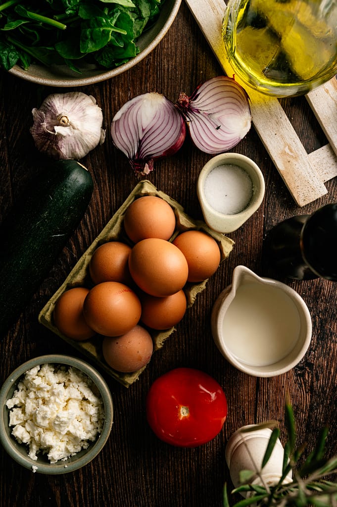 Ingredients for garden zucchini frittata arranged on a rustic table, including eggs, zucchini, tomato, spinach, onion, garlic, milk, cheese, and olive oil.