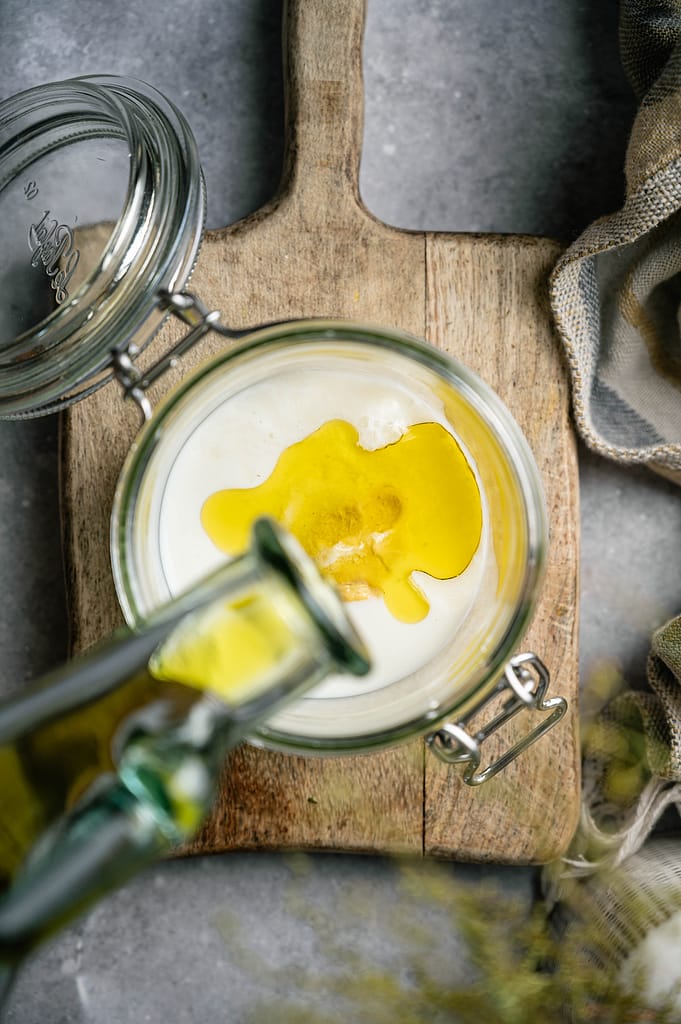 Oil being poured into pale ingredients in an open glass jar on a wooden board