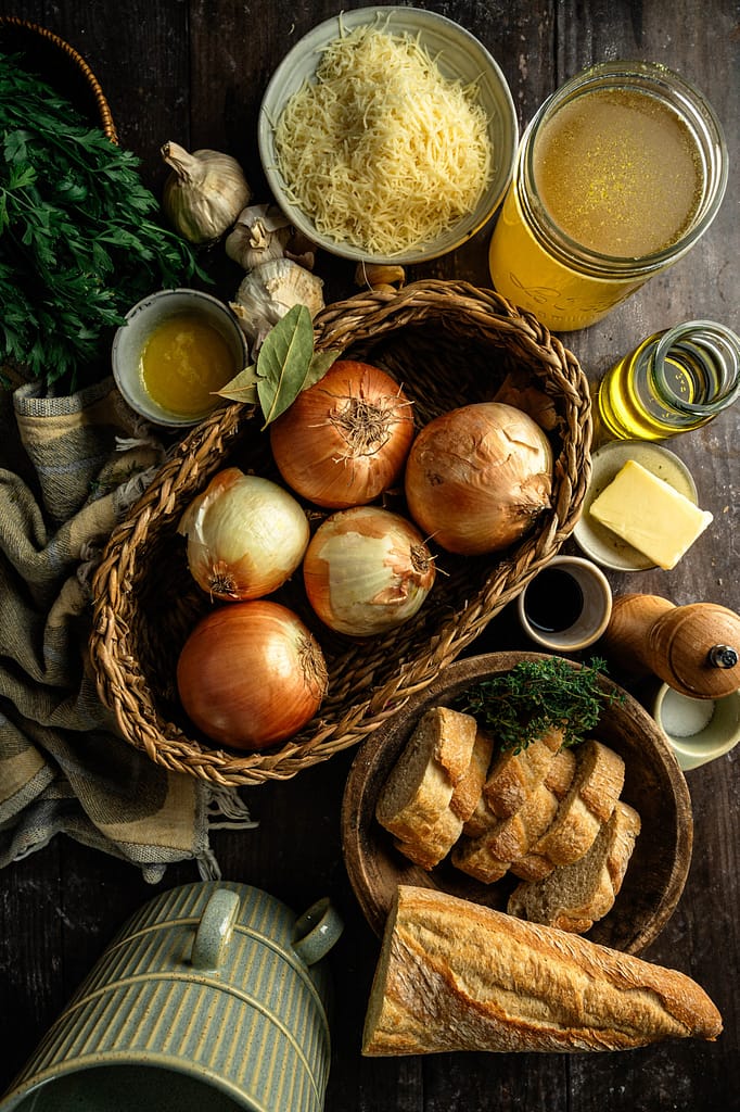 Yellow onions, grated cheese, broth, butter, olive oil, herbs, and sliced bread arranged on a dark wooden table.