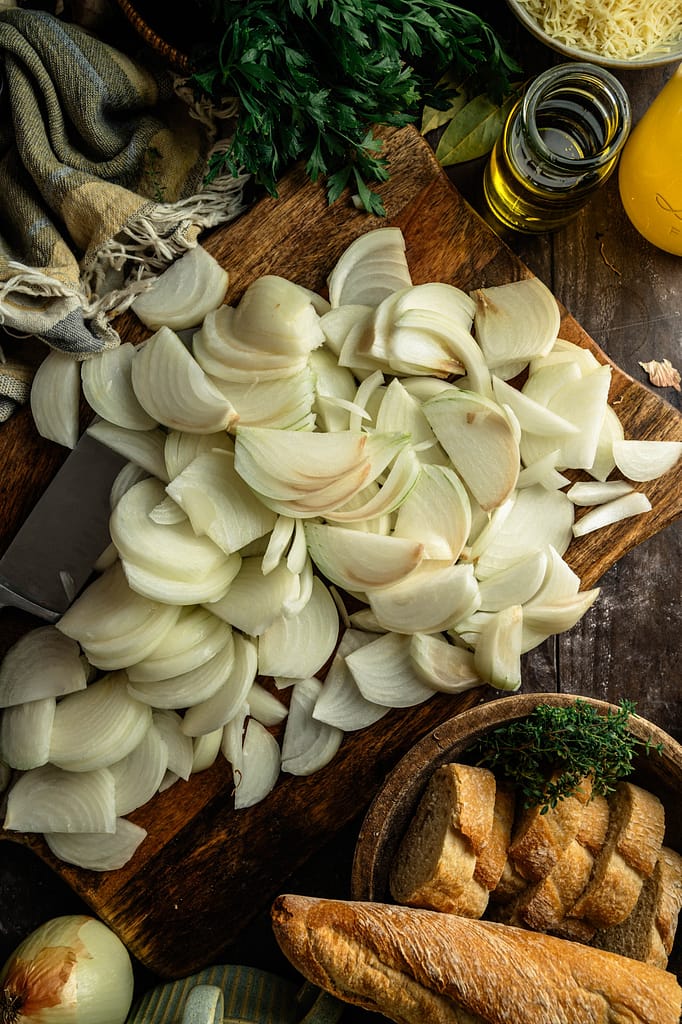 Sliced onions piled on a wooden cutting board beside a knife, herbs, olive oil, and bread.