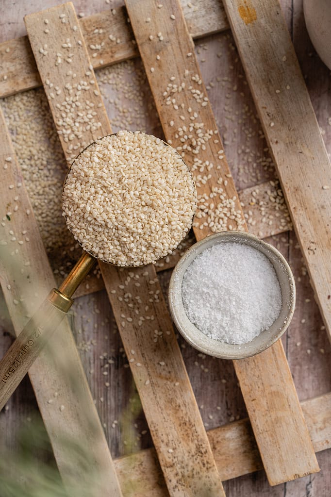 A measuring cup of sesame seeds beside a small bowl of salt on a wooden trivet.