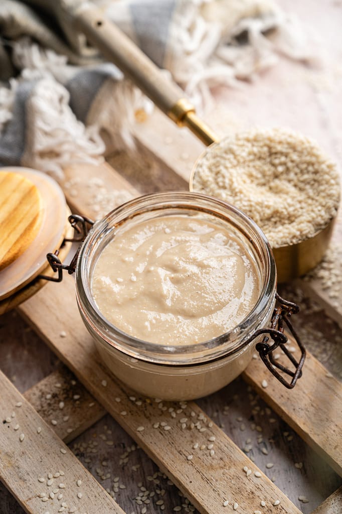 An open jar of homemade tahini with a wooden lid, scattered sesame seeds, and a filled measuring cup nearby.