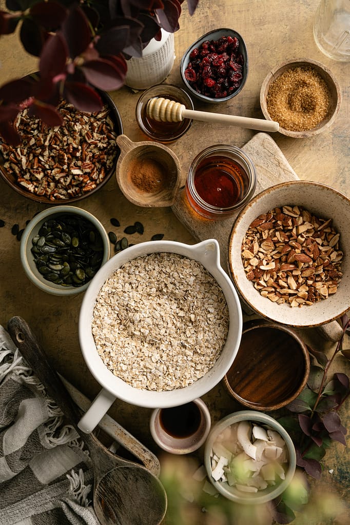 Bowls of oats, nuts, pumpkin seeds, coconut, dried cranberries, and sweeteners arranged for making granola.