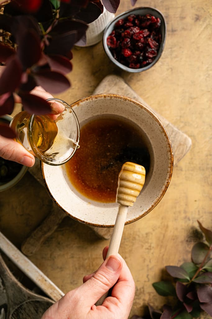 Coconut oil being poured into a bowl of maple syrup and honey beside a wooden honey dipper.