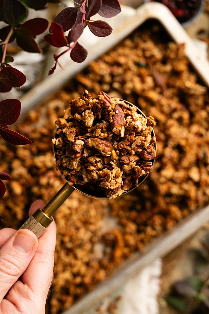 Close-up of a hand holding a measuring cup of golden granola clusters with nuts and cranberries.