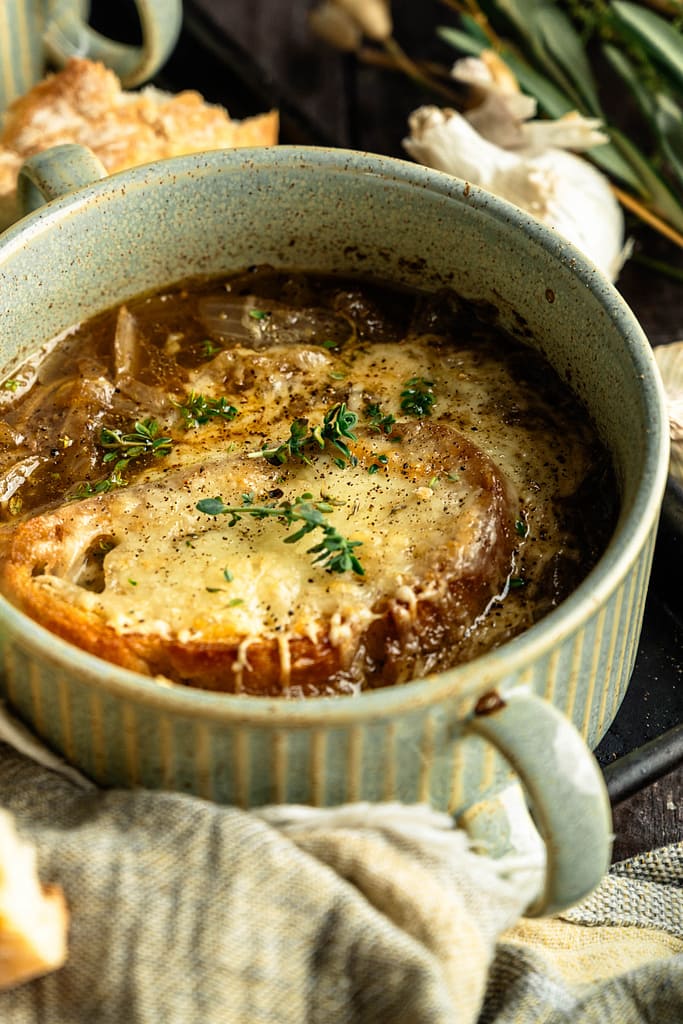 Close-up of golden cheese toast and thyme over French onion soup in a green handled crock.