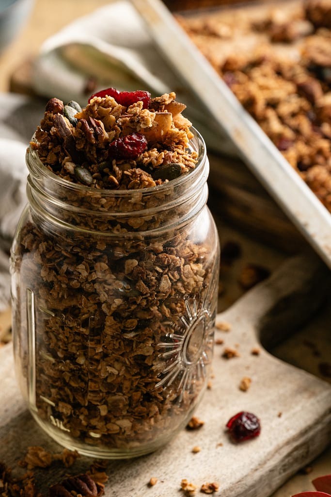 A glass jar filled to the rim with golden harvest granola, nuts, coconut, and dried cranberries.