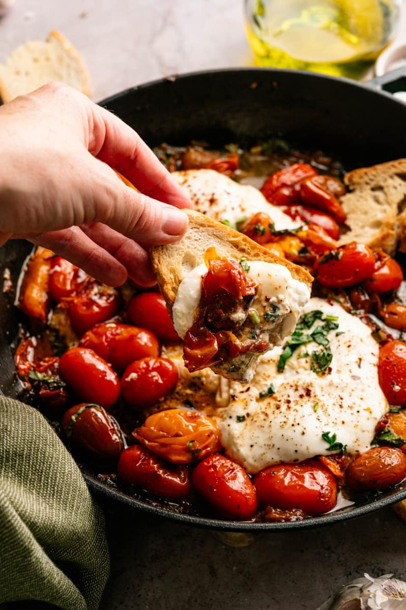 Hand dipping toasted artisan bread into warm creamy burrata topped with burst cherry tomatoes, garlic, basil, and olive oil in a rustic skillet.
