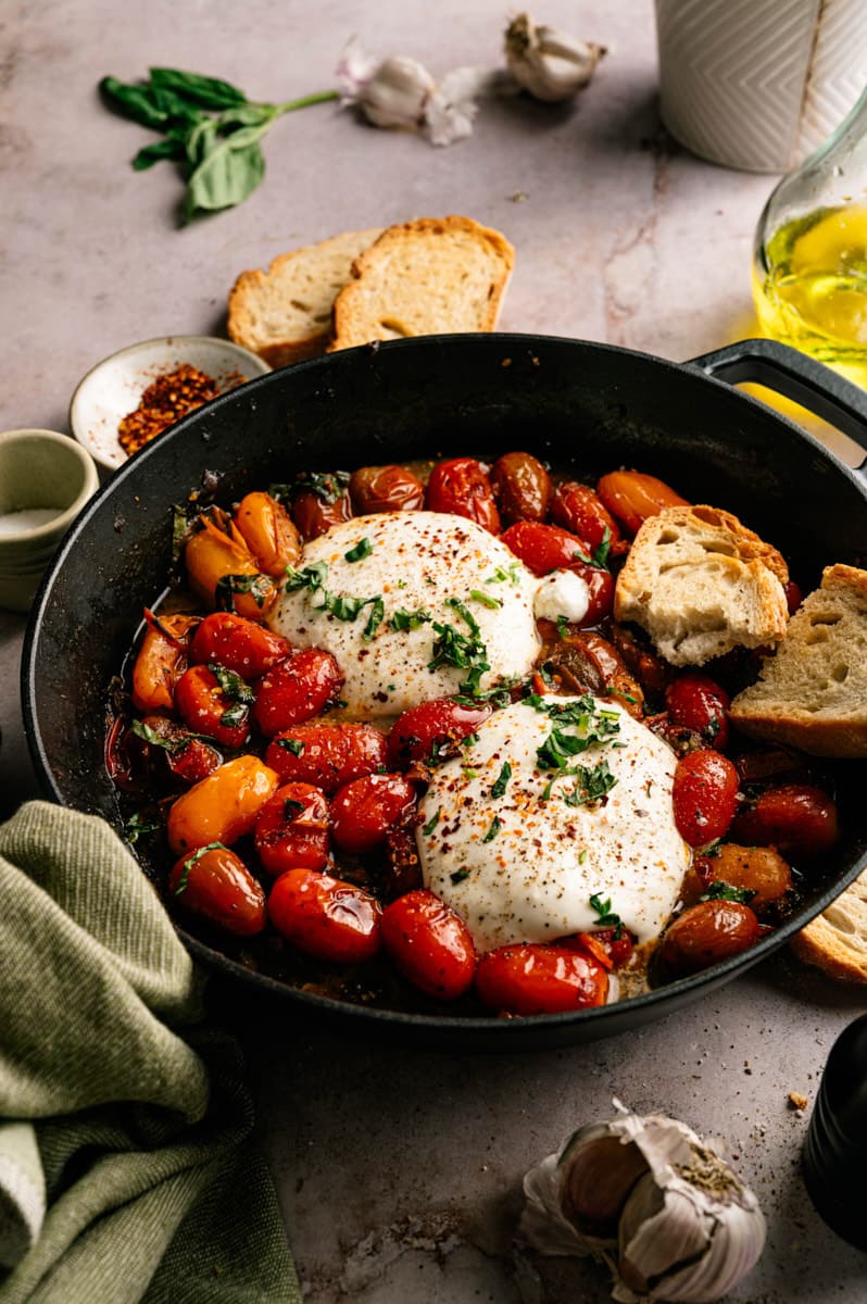 Rustic skillet filled with warm burrata, burst cherry tomatoes, garlic, basil, olive oil, and toasted artisan bread on a light stone surface.