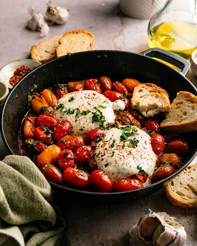 Warm burrata with colorful burst cherry tomatoes, basil, and rustic bread in a cast iron skillet.