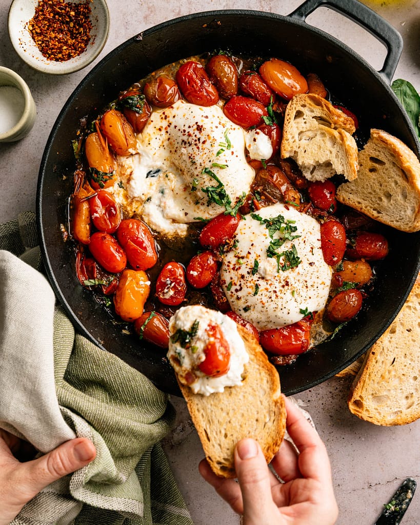 Overhead view of warm burrata and burst cherry tomatoes with a slice of rustic bread ready for serving.