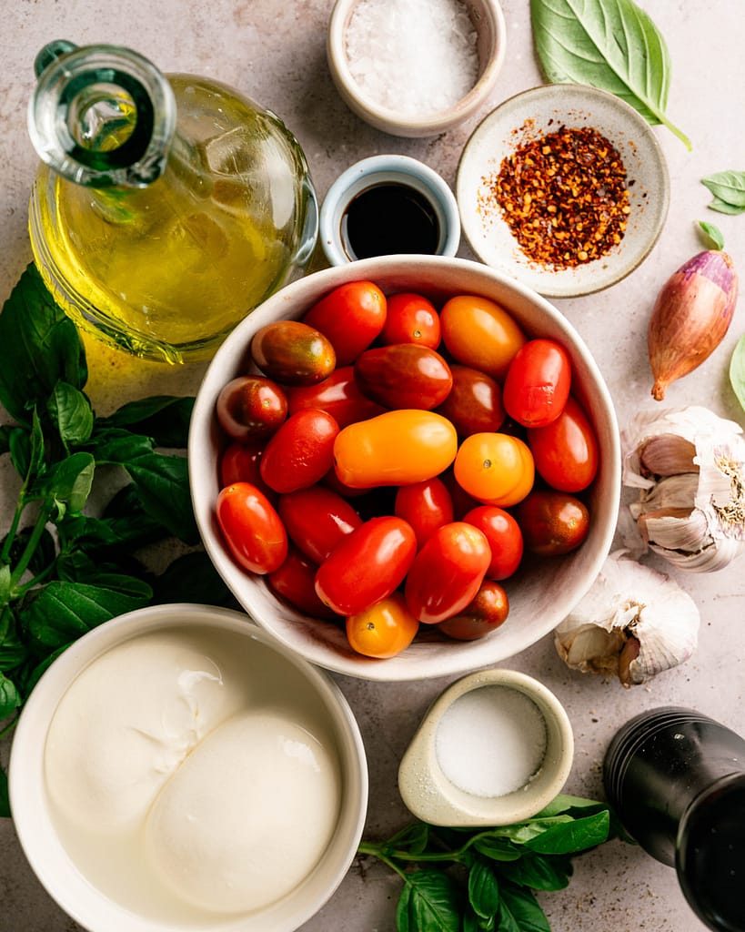 Fresh ingredients for warm burrata with burst cherry tomatoes, including burrata cheese, colorful cherry tomatoes, garlic, basil, olive oil, balsamic vinegar, sea salt, black pepper, and red pepper flakes arranged on a rustic surface.