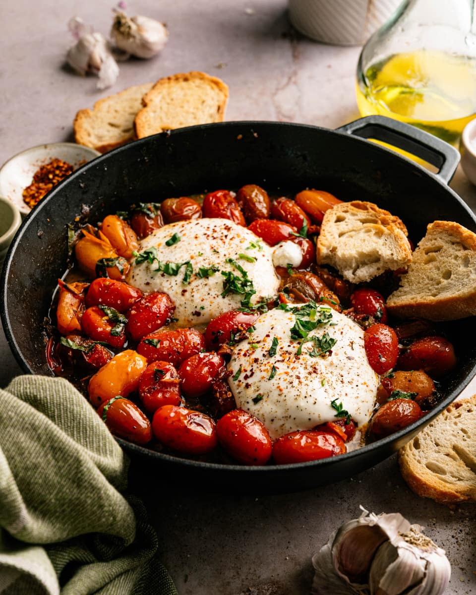 Warm burrata with colorful burst cherry tomatoes, basil, and rustic bread in a cast iron skillet.